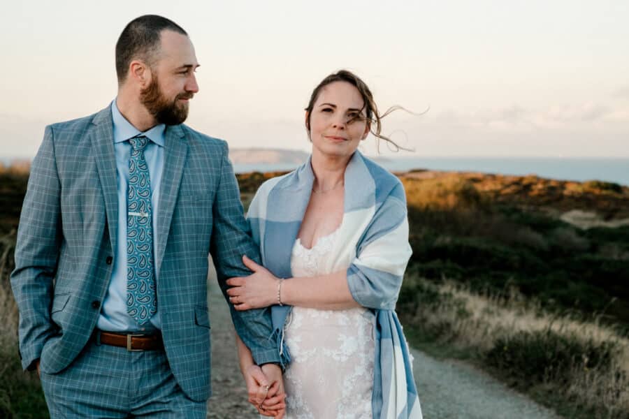 couple walking arm in arm in the Cliffs of Howth, groom looking loving at his bride as she stares at the camera, elopement in Howth