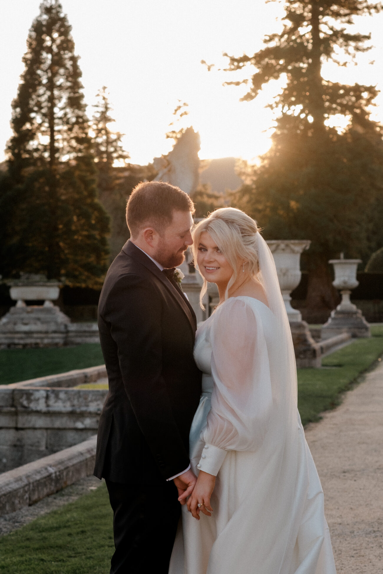 bride looking at the camera while the groom looks at the bride in the sunset at Powerscourt. wedding at Powerscourt estate 